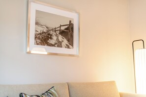 Une photographie encadrée représentant un paysage enneigé, accrochée au mur, un canapé avec un coussin et une lampe dotée d’un pied métallique.