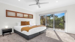 Un dormitorio moderno con una cama grande, un ventilador de techo, una mesita de noche y vistas a través de una puerta corrediza de cristal.