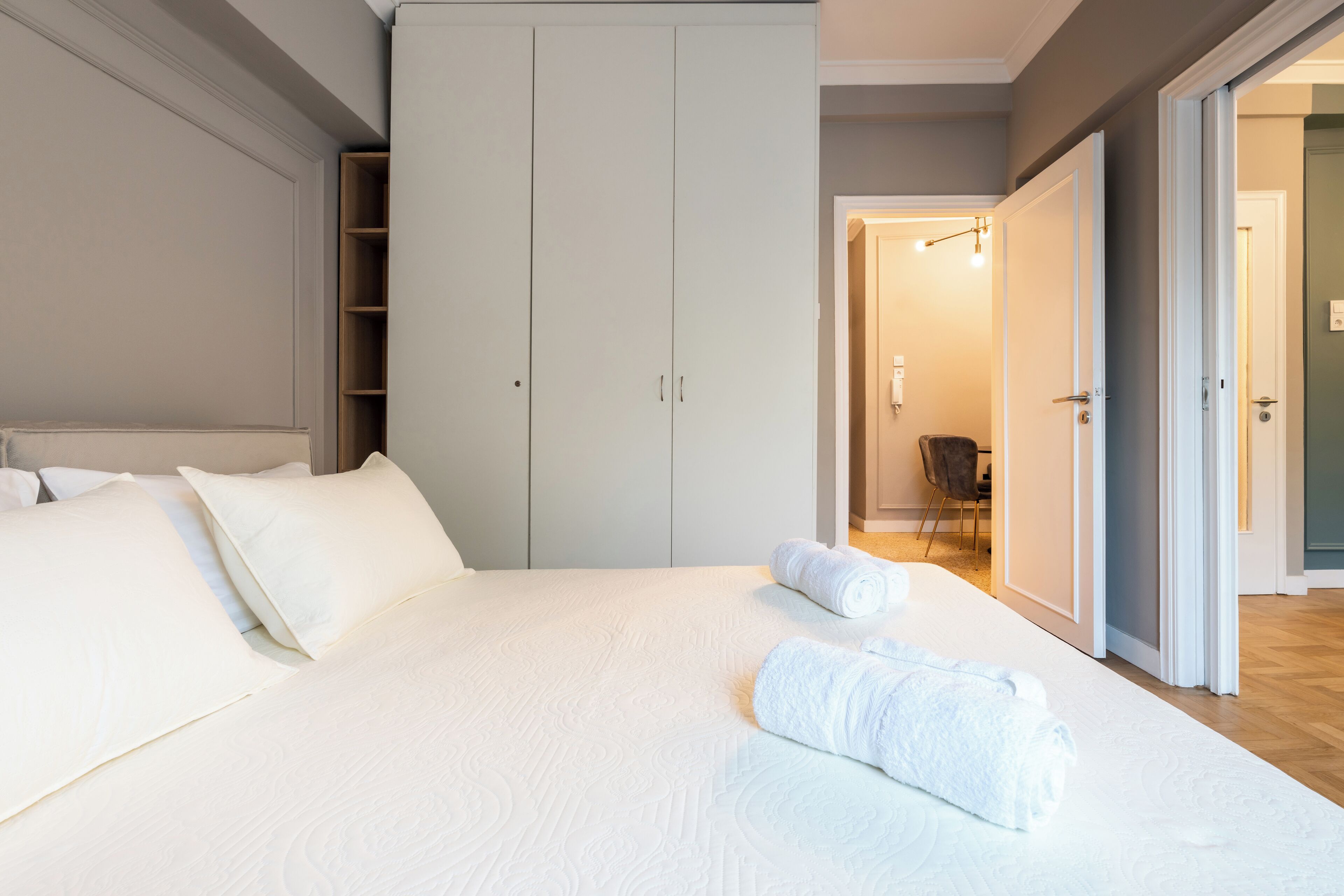 A neatly made bed with white linens and rolled towels, a large wardrobe, and a chair in the background.