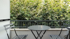 Een balkon met een ronde tafel en twee stoelen, omringd door een metalen leuning, met zicht op weelderige groene begroeiing op de achtergrond.
