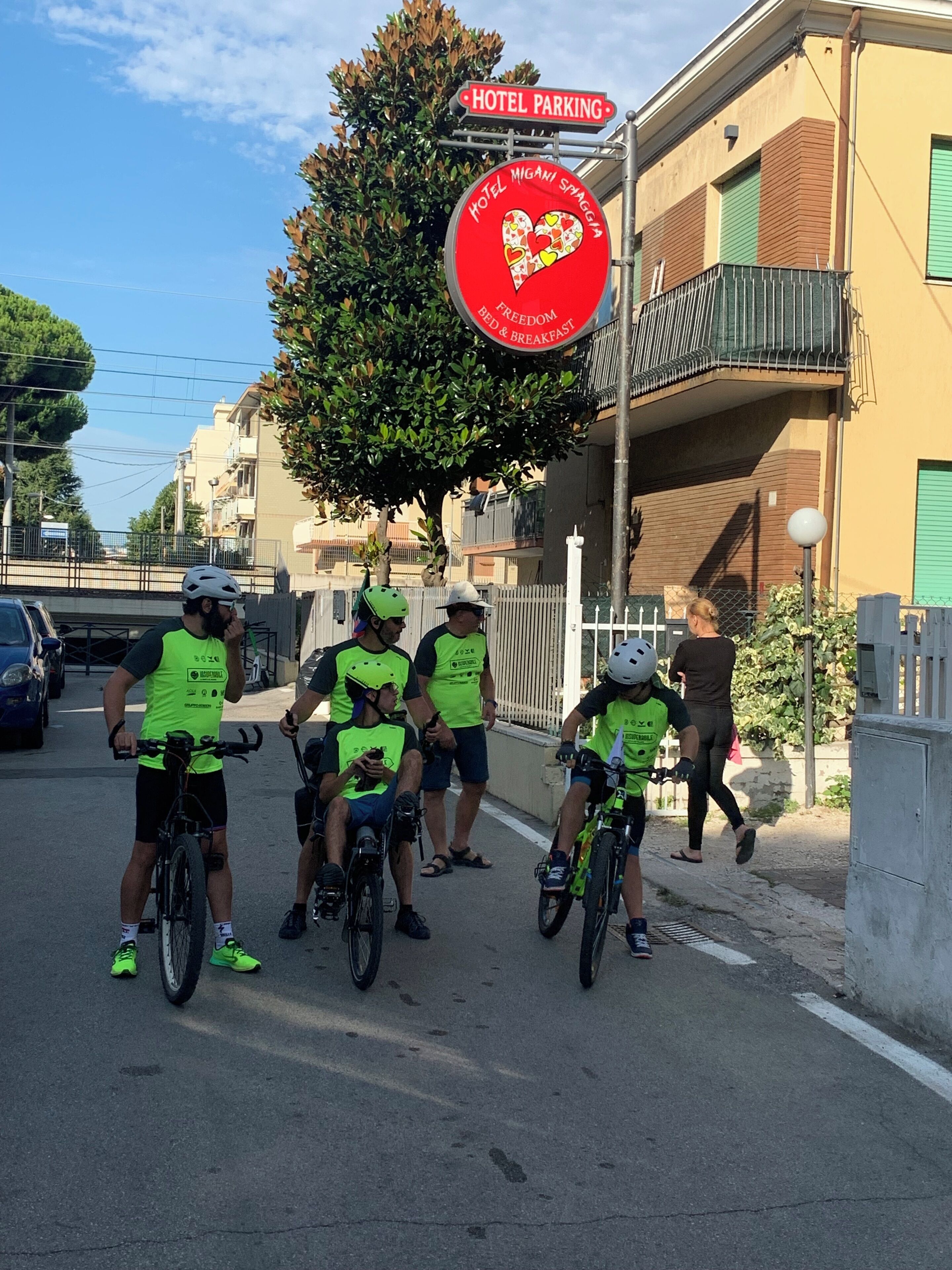 A group of cyclists in neon vests on a street with a hotel parking sign and a heart-shaped sign for Hotel Migani Spagna.
