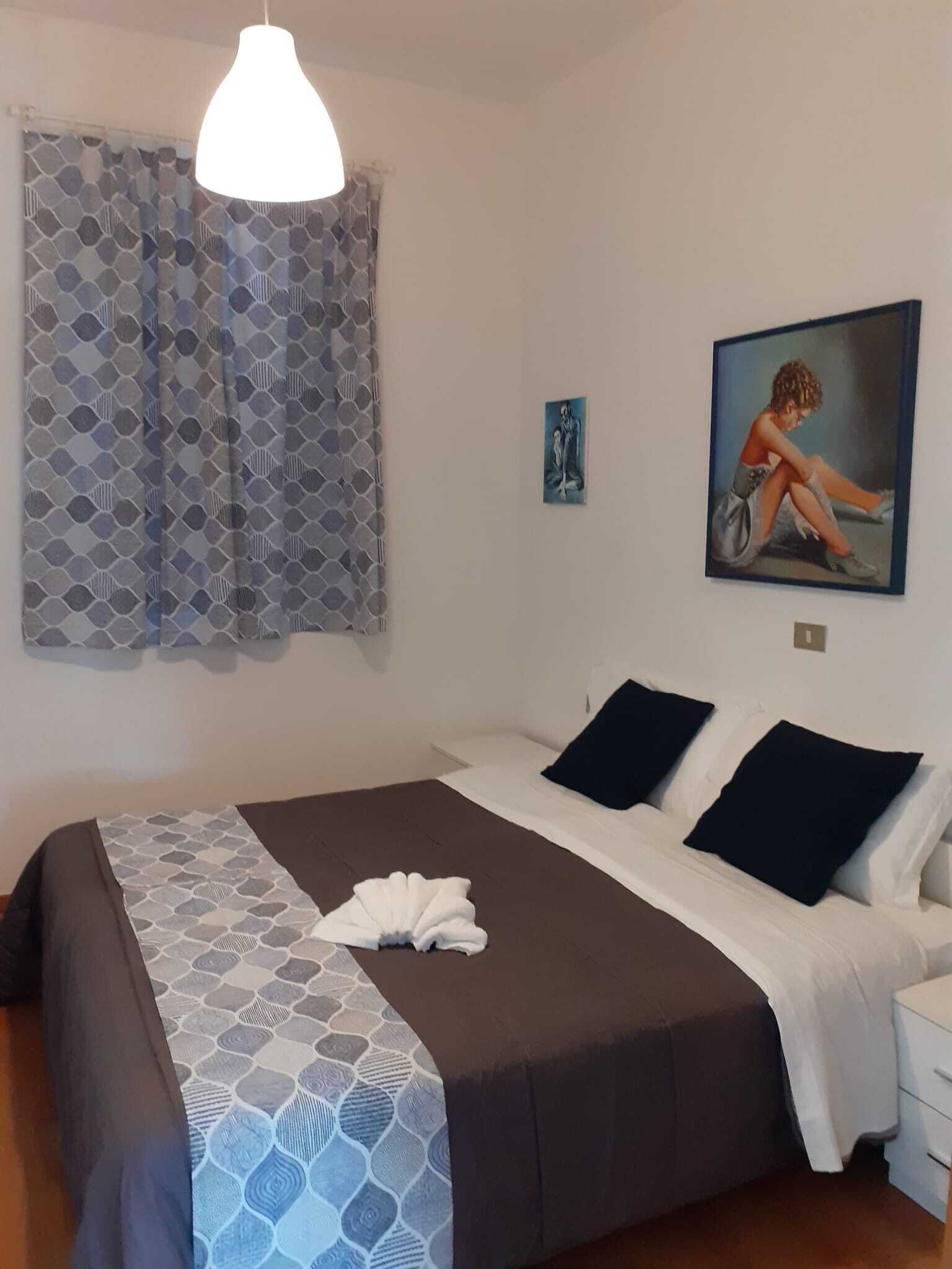 A bedroom with a bed, a nightstand, a painting, and a window with patterned curtains.