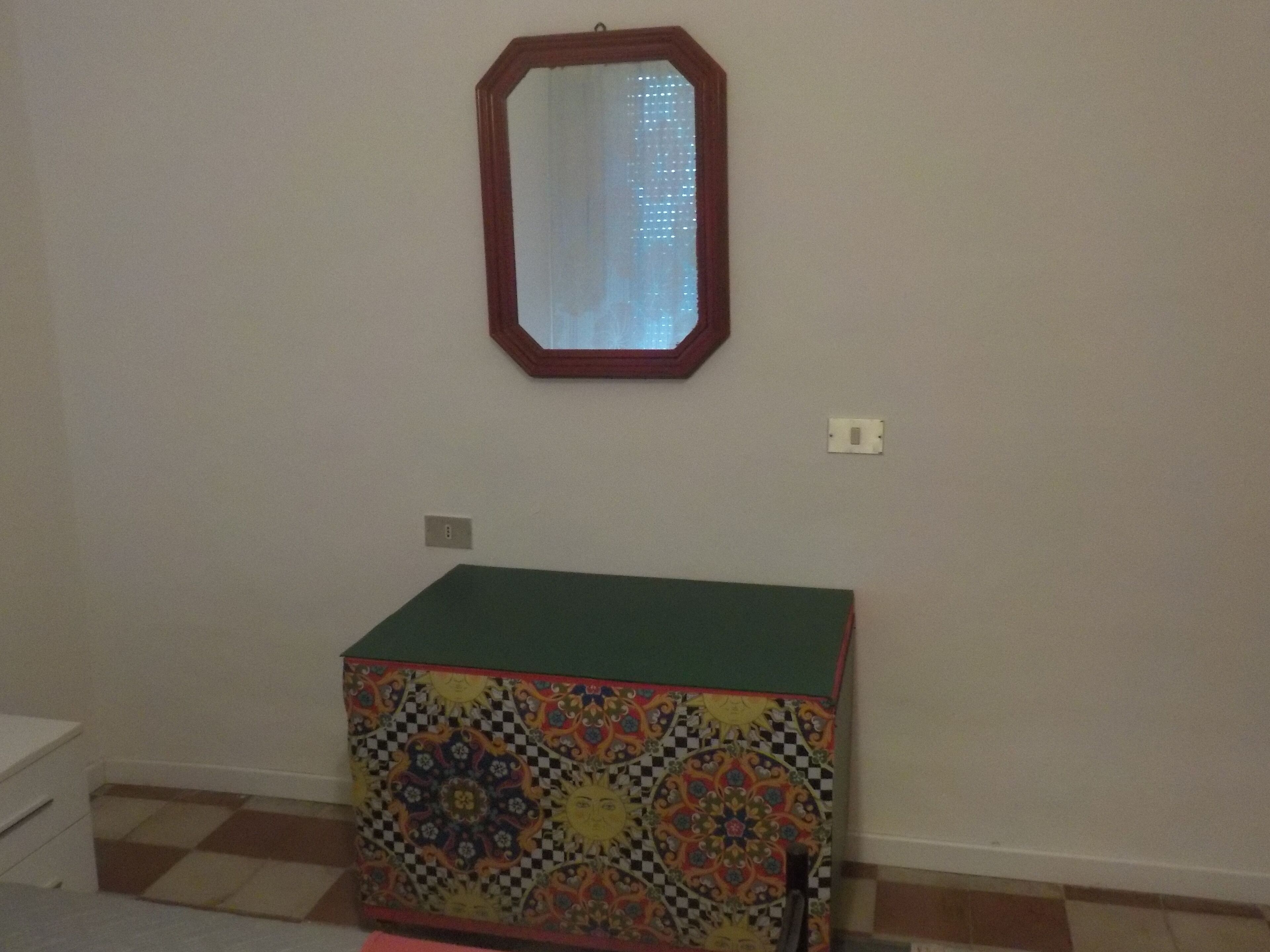 Een kamer met een klein, kleurrijk kastje met een groene bovenkant, een spiegel aan de muur en een betegelde vloer.