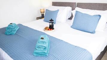 A neatly made bed with blue bedding, a small table with a coffee maker and snacks, and a lamp on a bedside table.