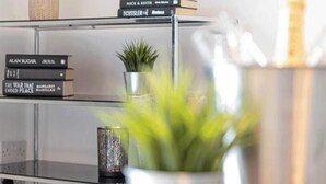 A modern interior with a glass table, a decorative plant, a metal vase, and a bookshelf with books. - Cozy Dundee Apartment With a View (Dundee)