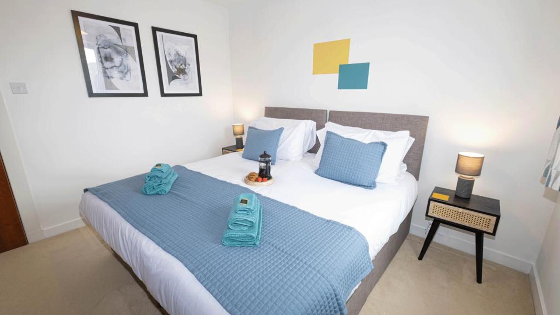 A neatly made bed with blue bedding and pillows, a bedside table with a lamp, and two framed pictures on the wall.