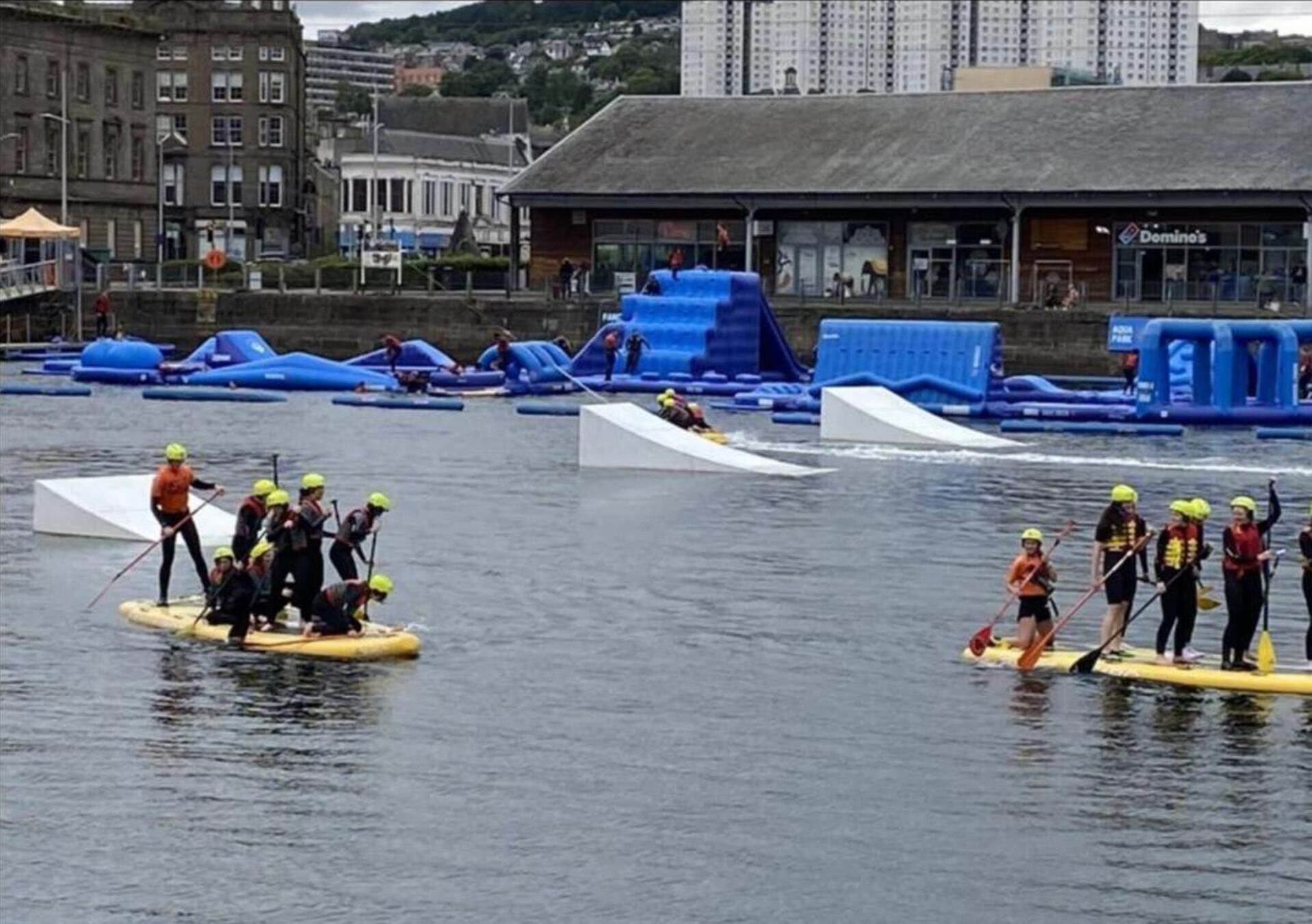 Een groep mensen op paddleboards in een watergebied, met gebouwen op de achtergrond.