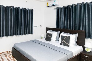 A neatly arranged hotel room with a large bed, dark curtains, a bedside table with a lamp, and an air conditioning unit on the wall. - Legit Luxury Apartments in Accra, Dome Pillar2 (Accra)