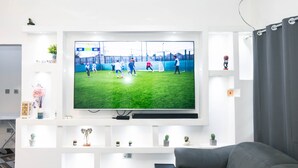 Un soggiorno con TV a parete che mostra una partita di calcio, un divano grigio e un tavolino da caffè in vetro.