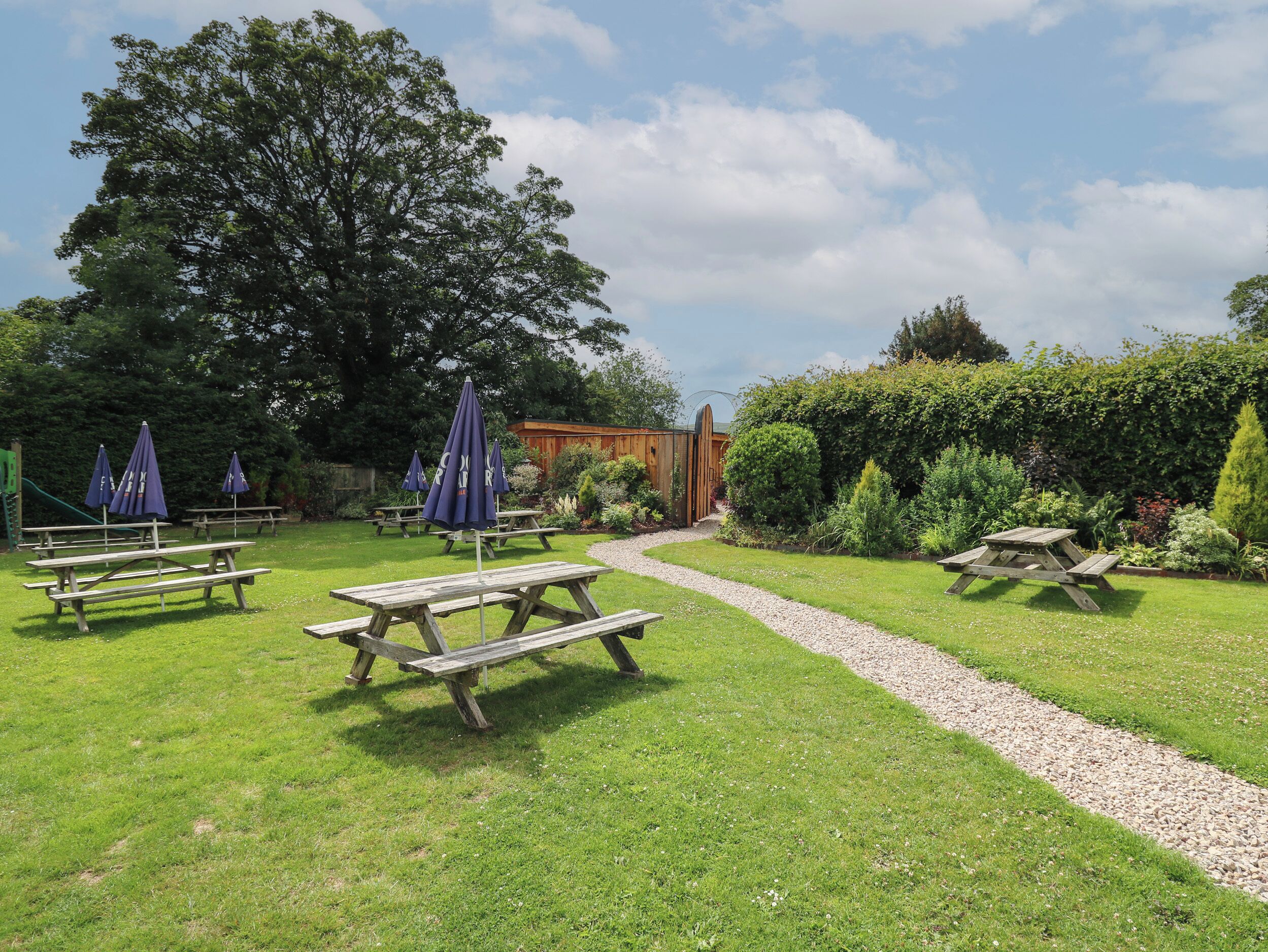 Un jardin avec des tables de pique-nique et des parasols, une clôture en bois et un chemin de gravier.