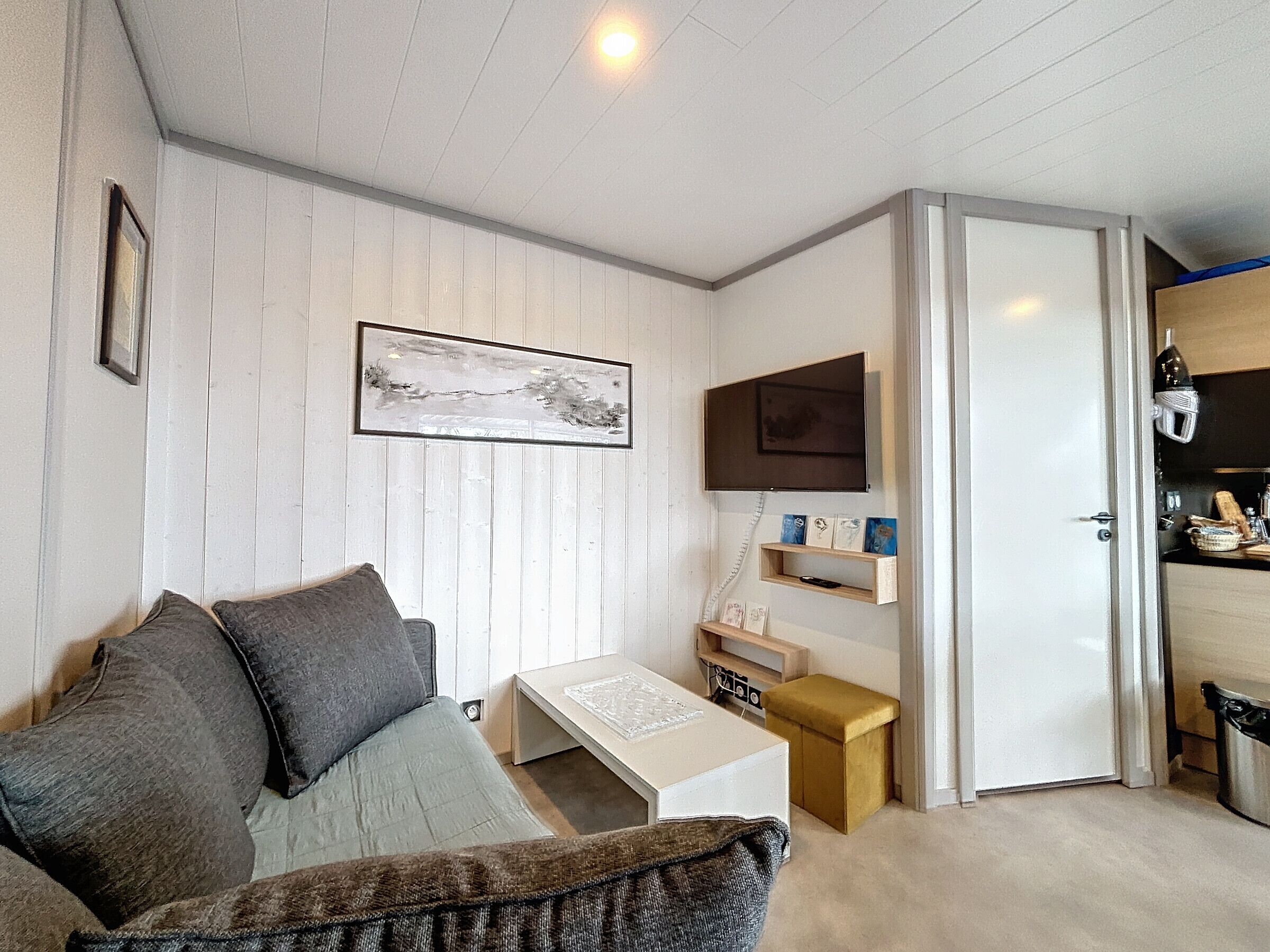 Un espacio habitable compacto con sofá, una mesita, televisor montado y una zona de cocina con microondas y estantes.