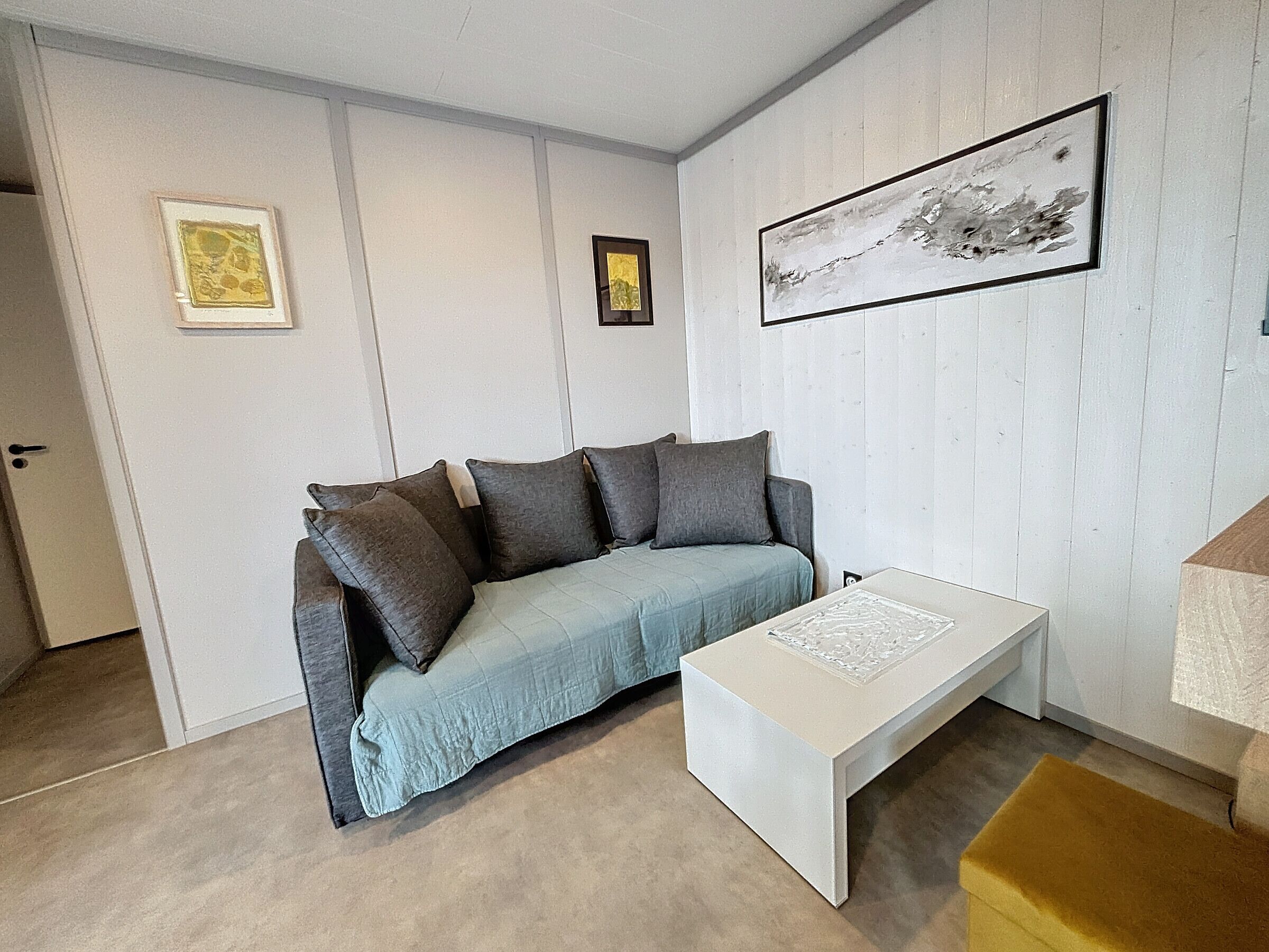 A room with a light blue sofa, a white coffee table, and a yellow ottoman. There are framed pictures on the walls.