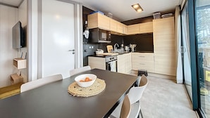 A modern kitchen with a dining area, featuring a dark countertop, white chairs, and a bowl of fruit. - Luxury Chalet - 4 Guests - Heated Pool - Close to Beach & Shops - Free WiFi (Jullouville)