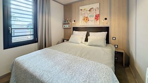 A modern bedroom with a large bed, a circular ceiling light, a window with blinds, and a wall-mounted shelf with decorative items. - Luxury Chalet - 4 Guests - Heated Pool - Close to Beach & Shops - Free WiFi (Jullouville)