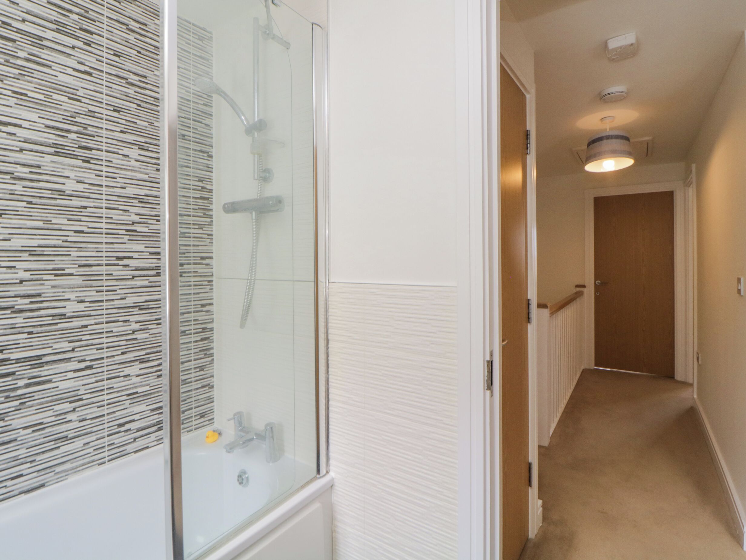 A hallway with a glass shower enclosure, a white tiled wall, and a wooden door at the end.