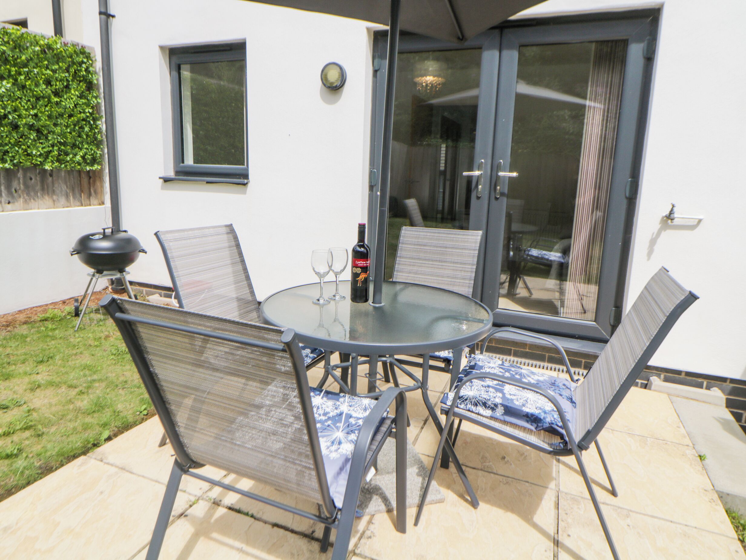 An outdoor patio with a round glass table, four chairs, a bottle of wine, and a barbecue grill.
