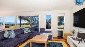 A modern living room with a sectional sofa, a coffee table, and a TV. Large windows offer views of the outdoors.