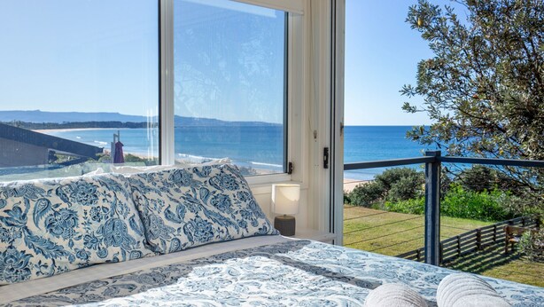 A bedroom with a large window overlooking a beach, a bed with patterned bedding, and a view of the ocean and distant landscape.