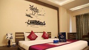 A hotel room with a bed, a nightstand, a wall decoration, a window with curtains, and a bedside table with a lamp and a bag. - Hotel Madhuvan Palace (Varanasi)