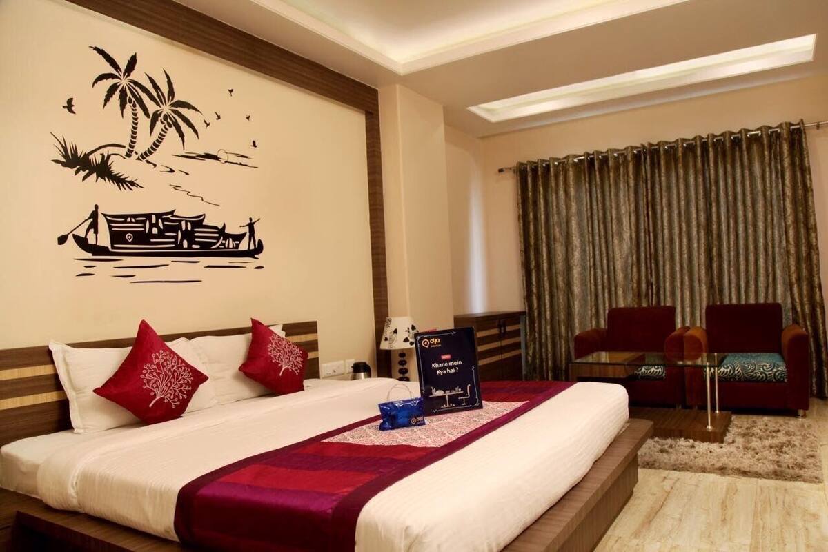 Una habitación de hotel con una cama, dos almohadas rojas, un mural en la pared, una mesita de cristal y dos sillones rojos, además de una cortina.