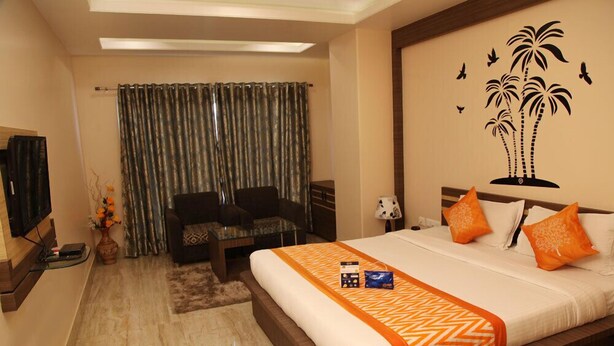A hotel room with a large bed, a desk, a chair, a TV, and a wall with a palm tree mural. - Hotel Madhuvan Palace (Varanasi)