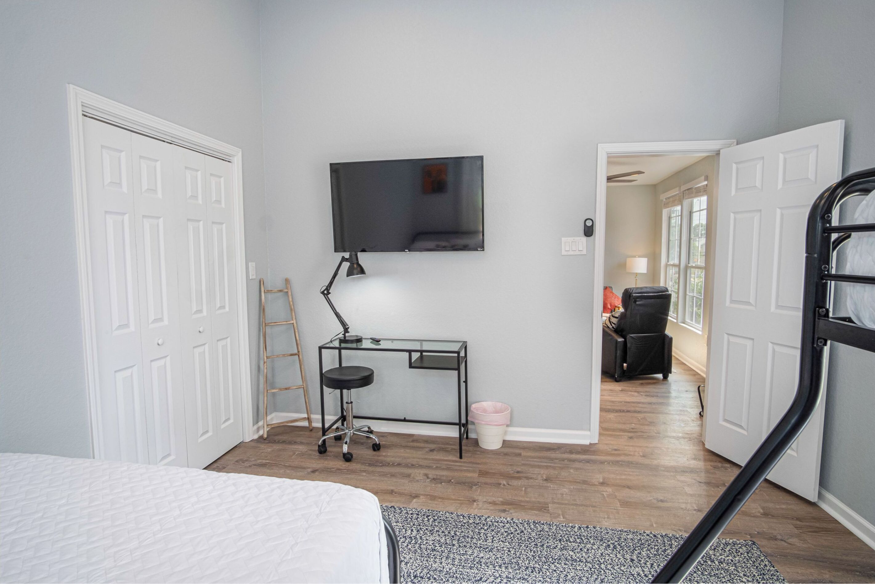 A bedroom with a bed, a desk with a chair, a TV mounted on the wall, and a door leading to another room.
