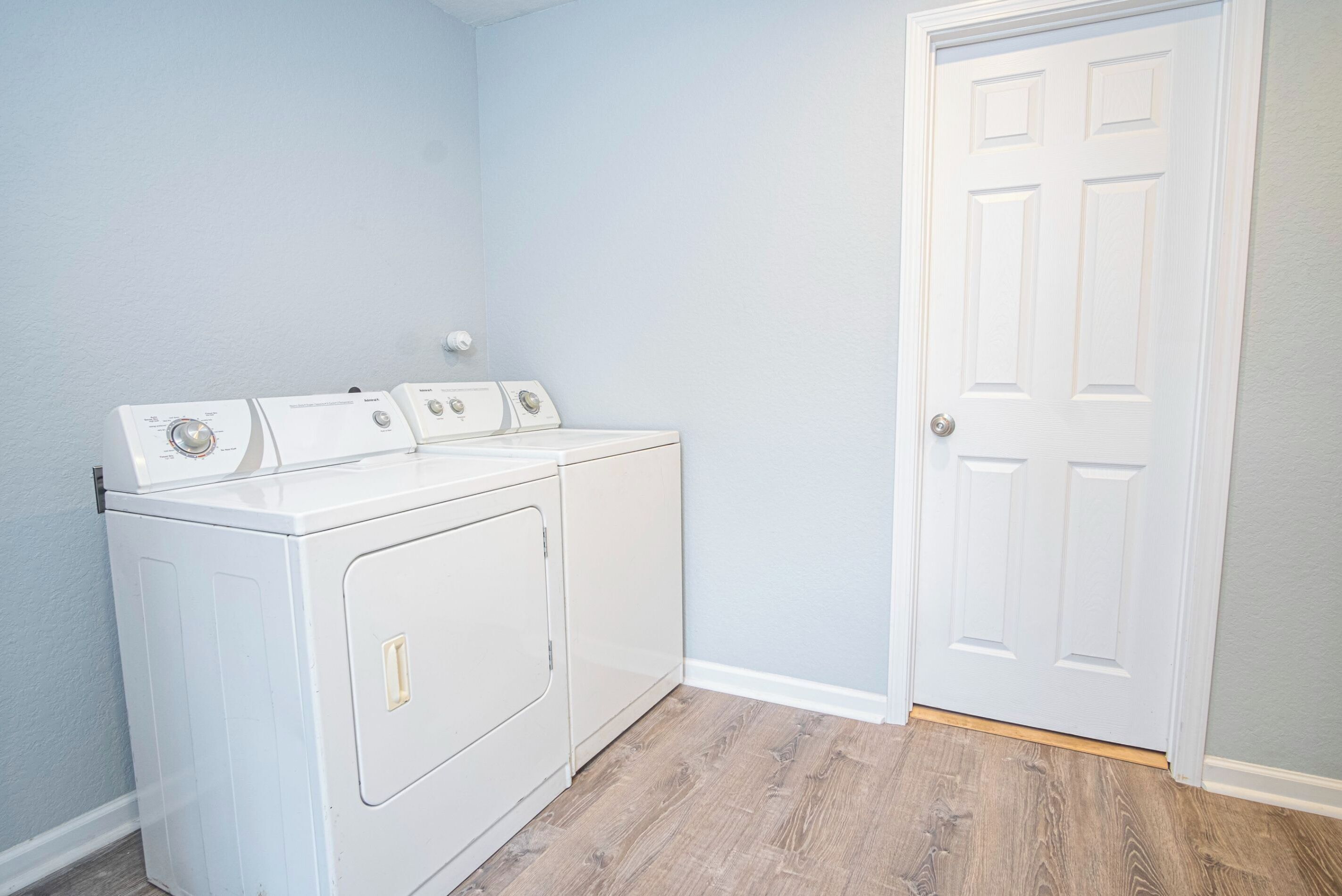 A laundry room with a front-loading washer and dryer, a wooden floor, and a white door.