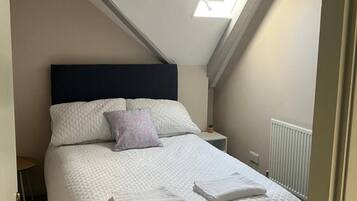 A bedroom with a bed, a headboard, a nightstand, a small table, a radiator, and a skylight.