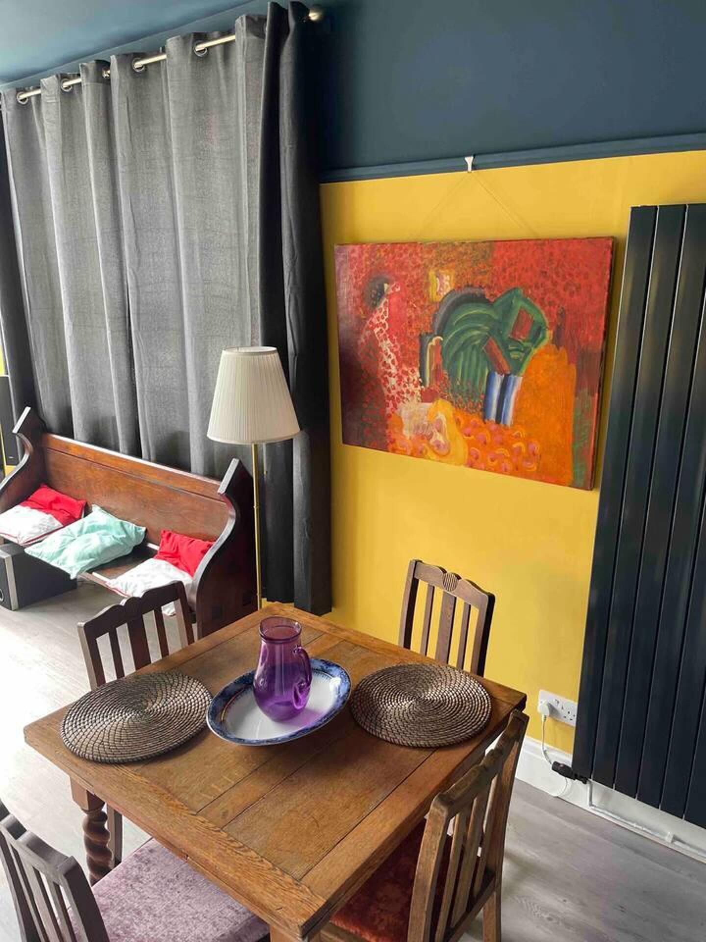 A dining area with a wooden table, chairs, a lamp, and a colorful abstract painting on the wall.