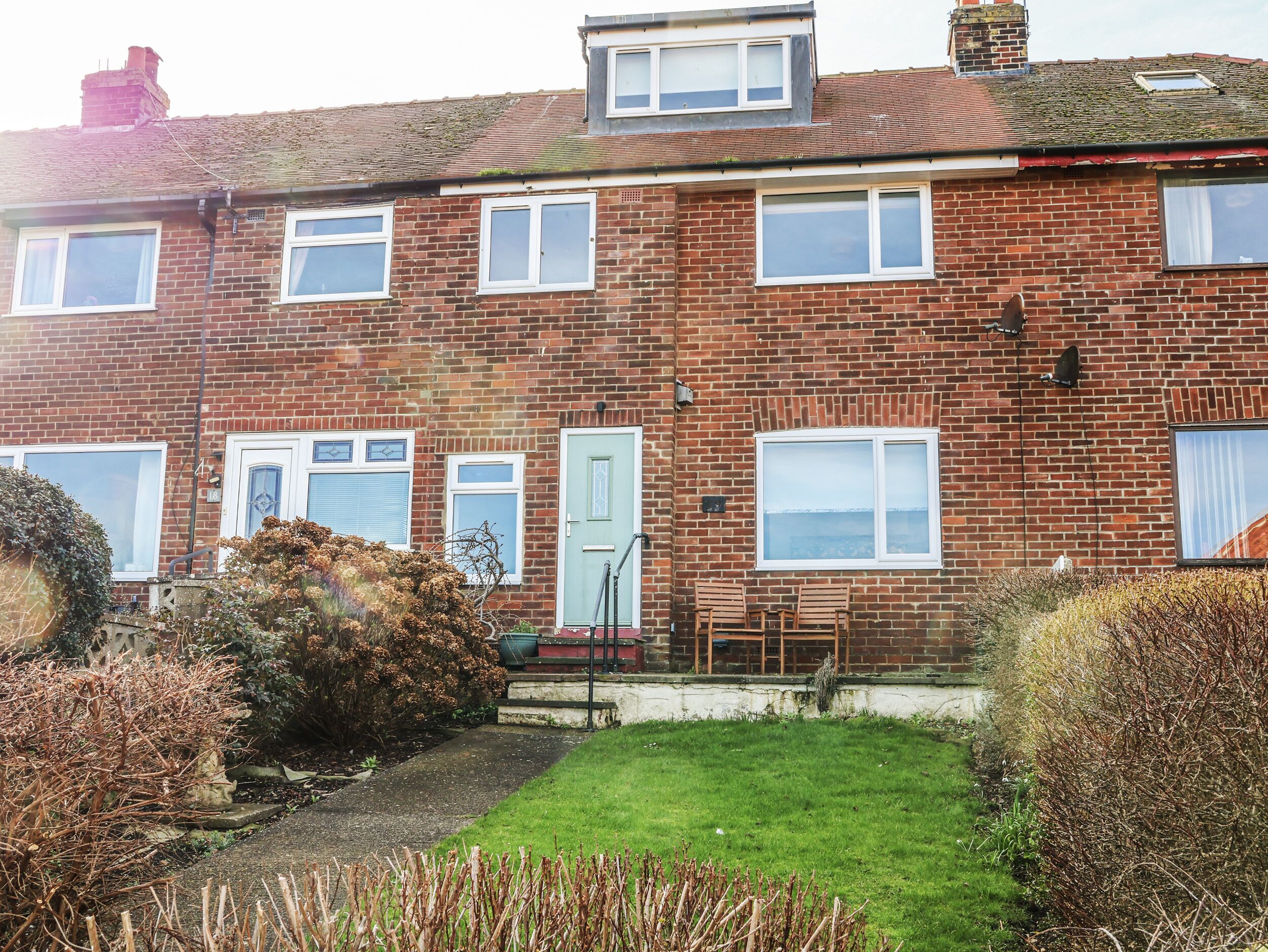 A two-story brick house with a front door, multiple windows, and a small garden area with a lawn and shrubs.