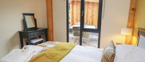 Un dormitorio con cama, mesita de noche con espejo, ventana con vistas al exterior y lámpara en la mesita auxiliar.