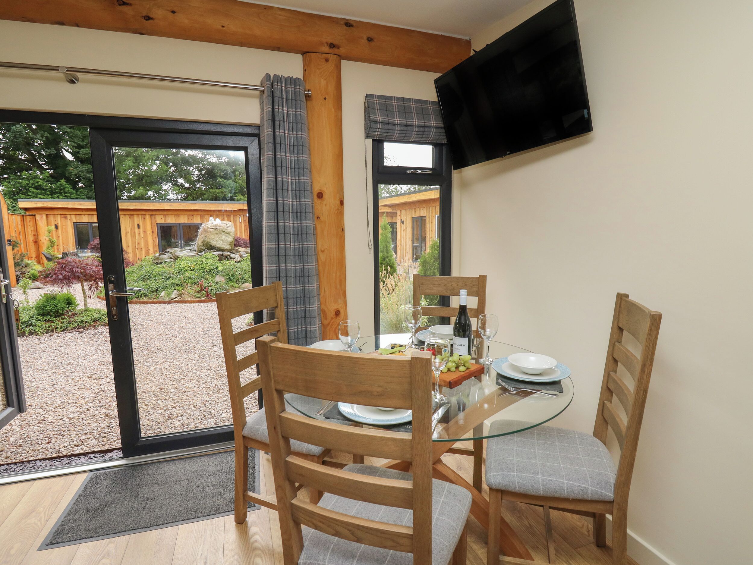 A dining area with a glass table, wooden chairs, a television mounted on the wall, and a view of a wooden building and garden through the glass door.