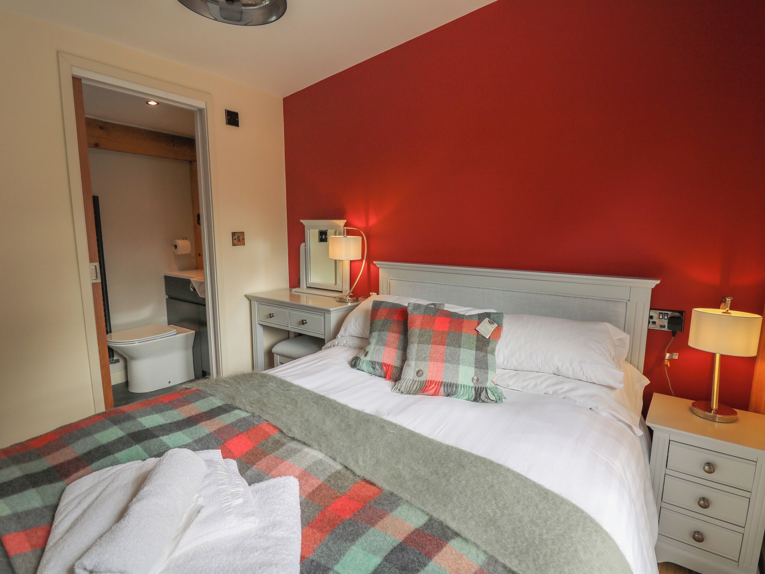 A hotel room with a bed, a nightstand, a bedside table, a mirror, a red wall, and a bathroom visible through an open door.