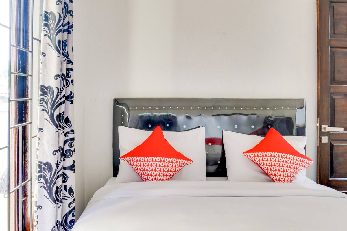 A bed with white bedding and two red pillows with white text, a headboard with a reflective surface, and a window with blue and white patterned curtains.