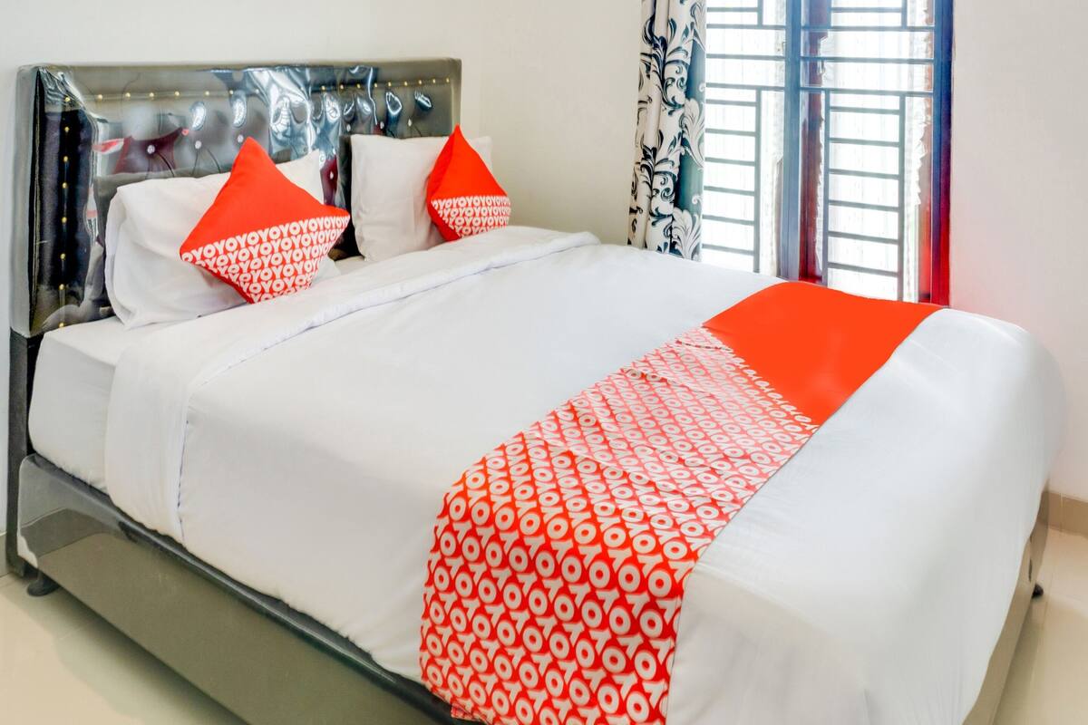 A neatly made bed with white and red bedding, a modern headboard, and a window with patterned curtains.