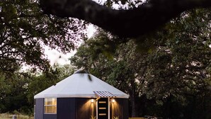 Una estructura tipo yurt con techo cónico, una puerta de madera y un pequeño porche con escalones que llevan a ella.