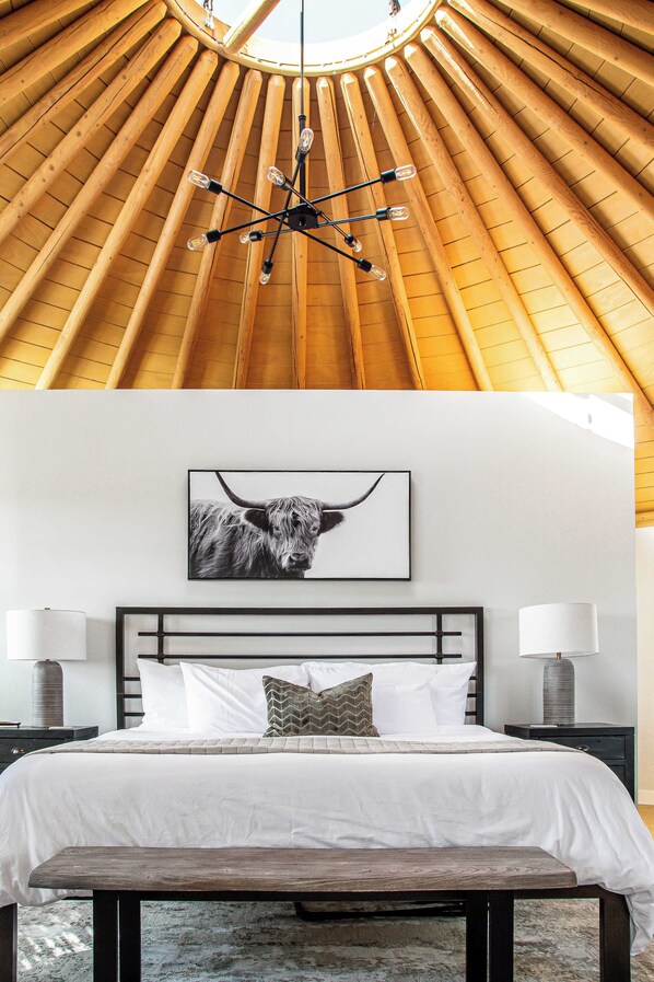 A bedroom with a wooden sloped ceiling, a bed with white bedding, a mounted artwork of a bull, bedside lamps, and a bench at the foot of the bed.