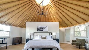 Un dormitorio con techo de madera inclinado, cama con ropa de cama blanca, un banco, una mesita de noche con lámparas y un ventanal grande.