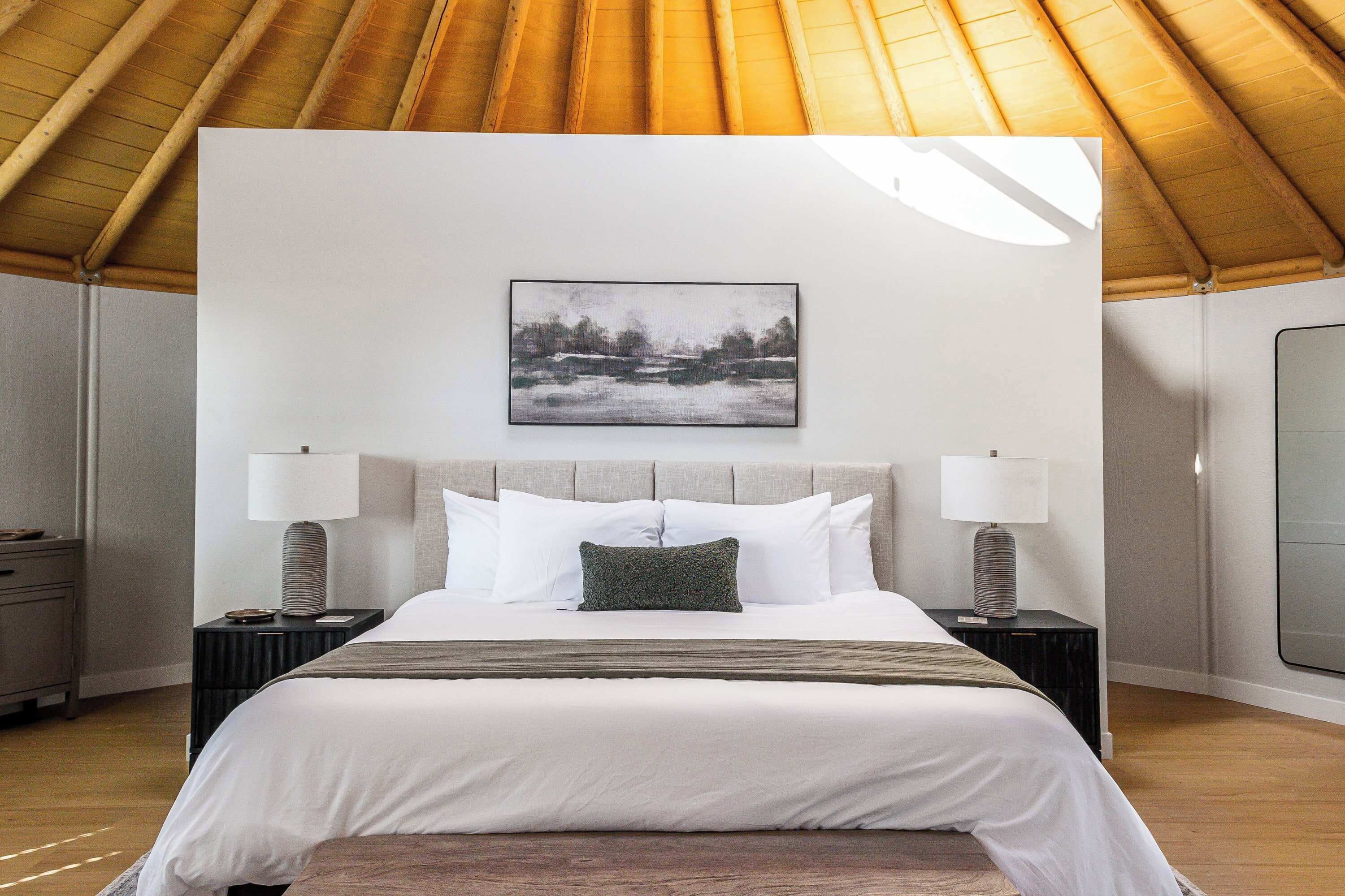 A bedroom with a large bed, two bedside tables with lamps, a framed picture on the wall, and a wooden ceiling.