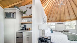 Een compacte keuken met een magnetron, koffiezetapparaat en een kleine koelkast. Grenzend aan de keuken bevindt zich een slaapkamer met een bed, nachtkastje en een uitzicht op de omgeving door grote ramen.
