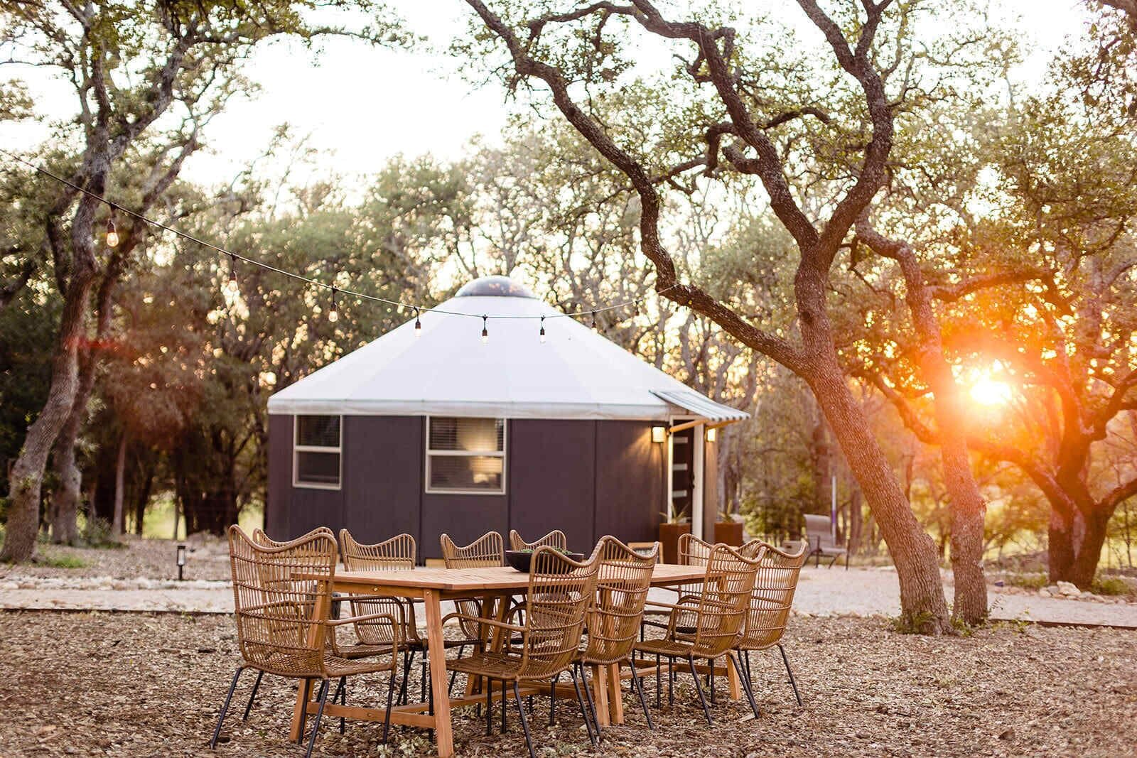 Een rustieke chalet met een puntdak, omringd door rieten stoelen en een tafel, gelegen in een bebost gebied met bomen en zonlicht dat erdoorheen schijnt.