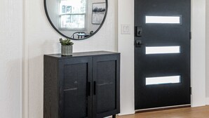 Een gang met een houten plafond, een zwarte deur met verlichte panelen, een ronde spiegel en een donkere houten kast met een plant.