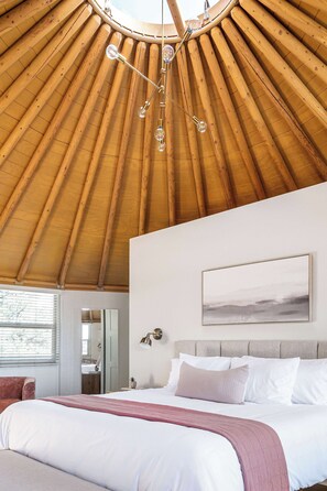 A bedroom with a wooden ceiling, a bed with white and pink bedding, a bedside table, and a wall-mounted light.