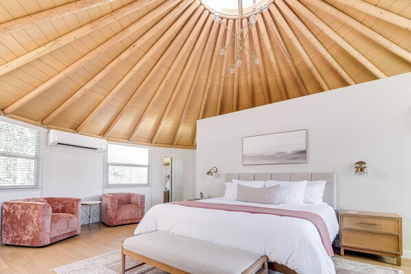 A bedroom with a wooden ceiling, a bed with white bedding, two red sofas, a bench, and a bedside table.