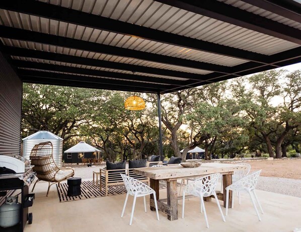 Un patio al aire libre con una mesa de madera y sillas blancas, rodeado de árboles y una zona de estar cubierta.