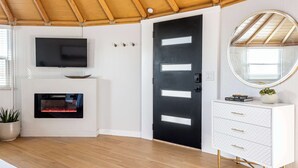 Ein modernes Interieur mit Holzbalkendecke, einer schwarzen Tür mit weißen Füllungen, einer weißen Konsole mit Schubladen, einem Flachbildfernseher über einem Kamin und einem runden Spiegel auf einer Konsole.