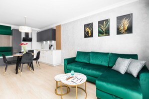 A modern living room with a green sofa, a dining table with chairs, a kitchen area, and three framed artworks on the wall.