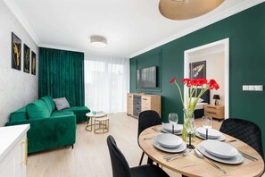 A modern living room with a green sofa, a dining table set for four, and a TV on the wall.