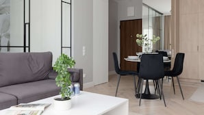 Un salon moderne avec un canapé gris, une table basse blanche sur laquelle sont posés des magazines, une plante en pot et un coin repas avec une table en bois et des chaises noires.
