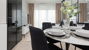 Un espace repas moderne avec une table ronde pour quatre personnes, des chaises noires à revêtement en tissu, un vase avec des plantes vertes et une grande fenêtre ornée de rideaux légers.